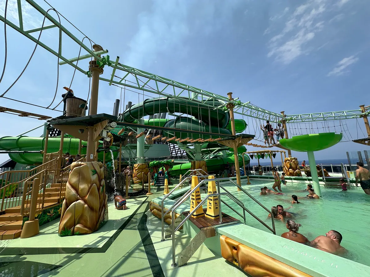 People enjoying a water park with green slides, a rope course, and a pool under a clear blue sky.
