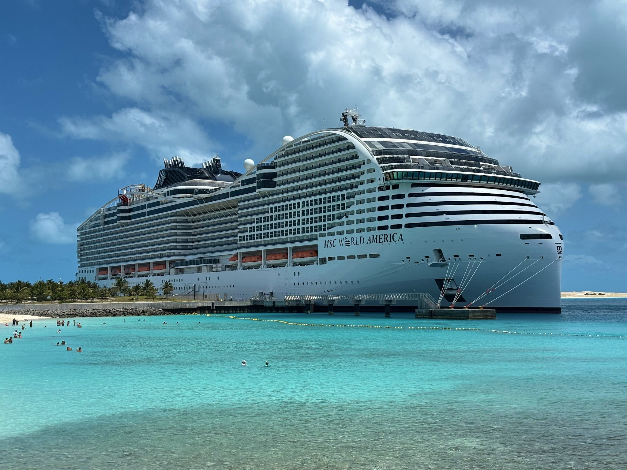 A large cruise ship MSC World America docked near a tropical beach with turquoise water and a few swimmers under a partly cloudy sky.