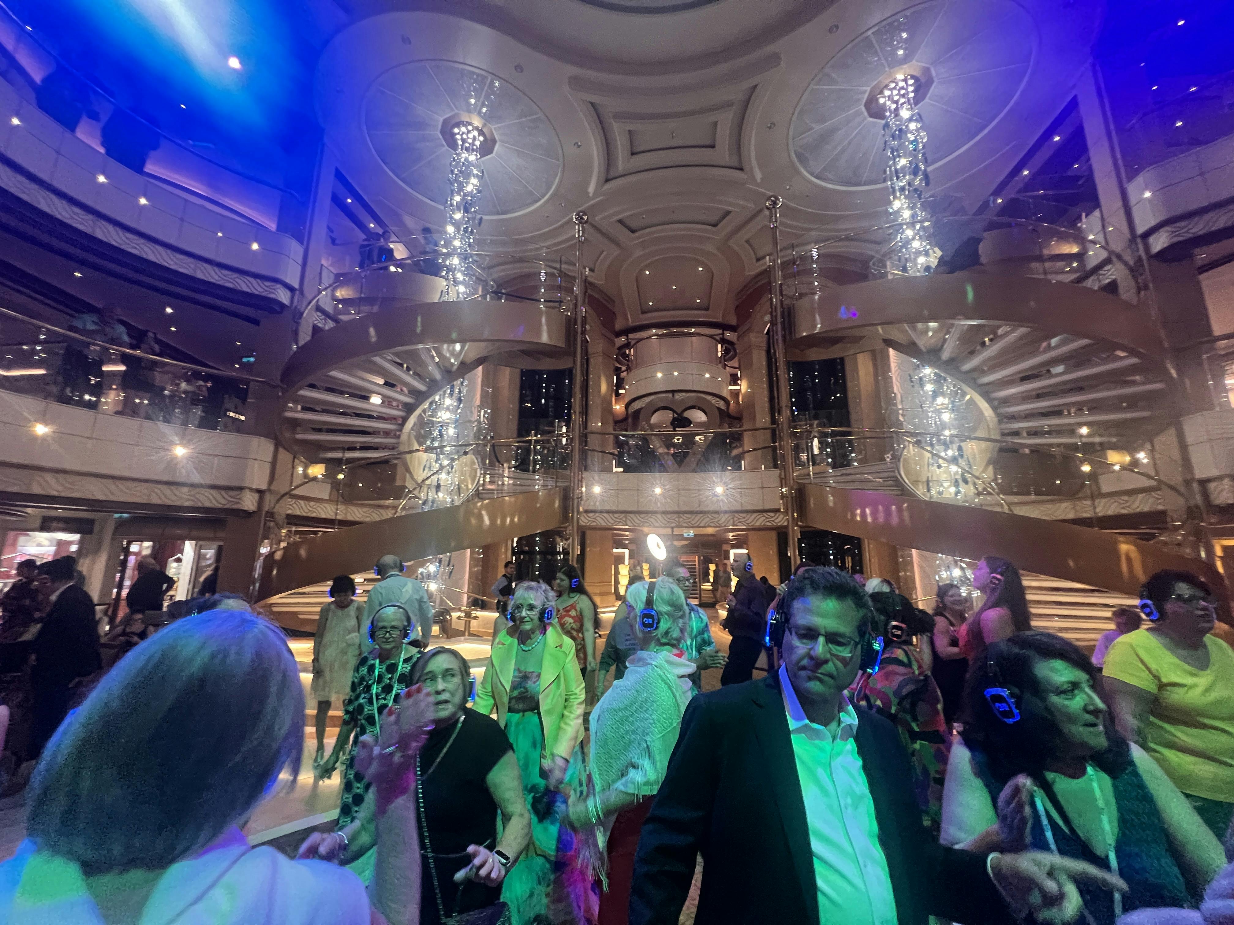 People dance in a cruise ship atrium with spiral staircases and dramatic lighting.