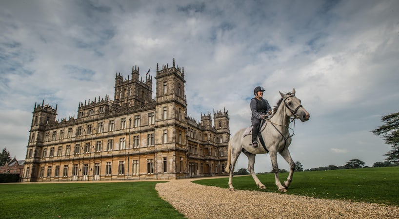 Moody exterior shot of a woman riding a light grey horse in front of Highclere Castle