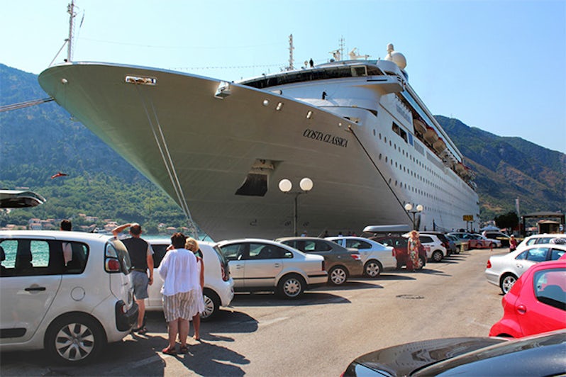 people walk on the pier to which moored cruise ship in a port surrounded by mountains. along the pier there are numerous cars