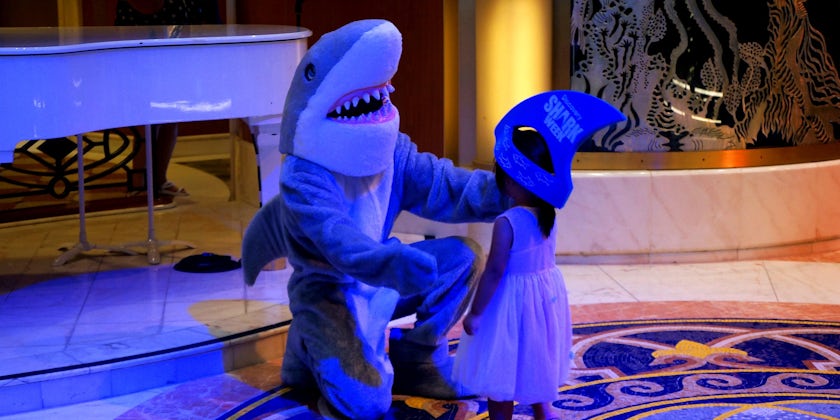A Princess Cruises crewmember in a shark costume, posing with a little girl wearing a Shark Week hat
