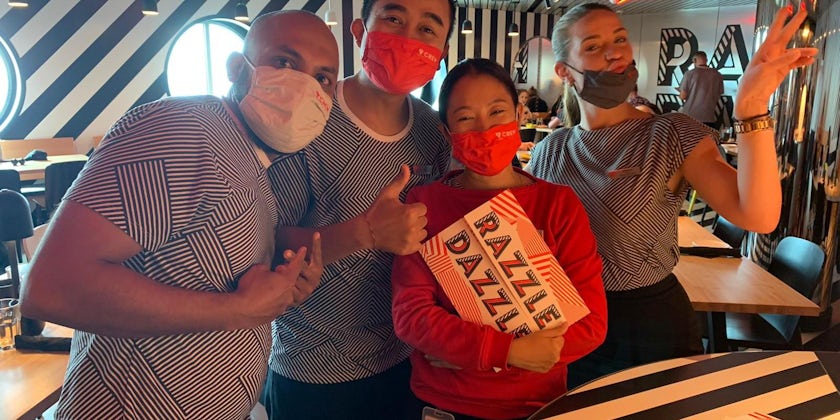 Crew from Razzle Dazzle pose with menu and masks in restaurant on Scarlet Lady (Photo: Adam Coulter)